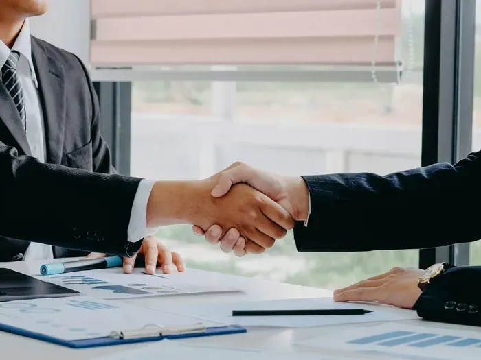 Two business professionals in suits shaking hands across a desk with charts and documents in bright office